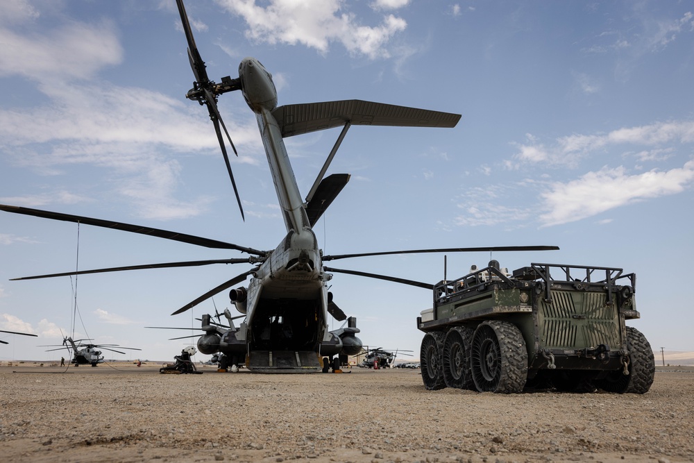 A large military heavy-lift helicopter, consistent with a Sikorsky CH-53E Super Stallion, is positioned on an arid desert airfield with its rear cargo ramp open. Adjacent to the helicopter is a multi-wheeled military ground vehicle, possibly an autonomous or remote-controlled logistics platform. Several additional helicopters are visible in the background on what appears to be a forward operating base or training area.