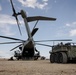 A large military heavy-lift helicopter, consistent with a Sikorsky CH-53E Super Stallion, is positioned on an arid desert airfield with its rear cargo ramp open. Adjacent to the helicopter is a multi-wheeled military ground vehicle, possibly an autonomous or remote-controlled logistics platform. Several additional helicopters are visible in the background on what appears to be a forward operating base or training area.