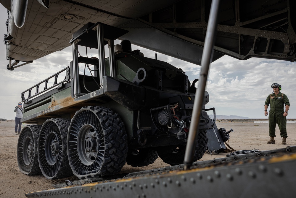 A military ground vehicle with distinctive airless/non-pneumatic wheels is being loaded or unloaded from a large military transport aircraft via a rear cargo ramp. The scene takes place on an arid desert airstrip, with at least two personnel visible overseeing the operation. The vehicle appears to be a multi-wheeled autonomous or semi-autonomous military utility platform.
