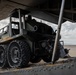 A military ground vehicle with distinctive airless/non-pneumatic wheels is being loaded or unloaded from a large military transport aircraft via a rear cargo ramp. The scene takes place on an arid desert airstrip, with at least two personnel visible overseeing the operation. The vehicle appears to be a multi-wheeled autonomous or semi-autonomous military utility platform.