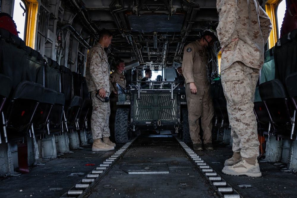 The image shows the interior of a large military transport helicopter, likely a CH-46 Sea Knight or CH-53, with personnel loading or securing a military utility vehicle. Several individuals in U.S. Marine Corps MARPAT desert camouflage uniforms and tan flight suits are visible overseeing the cargo operation. The cargo bay features fold-down troop seats along both sides and a center floor track system for securing vehicles and equipment.