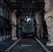 The image shows the interior of a large military transport helicopter, likely a CH-46 Sea Knight or CH-53, with personnel loading or securing a military utility vehicle. Several individuals in U.S. Marine Corps MARPAT desert camouflage uniforms and tan flight suits are visible overseeing the cargo operation. The cargo bay features fold-down troop seats along both sides and a center floor track system for securing vehicles and equipment.