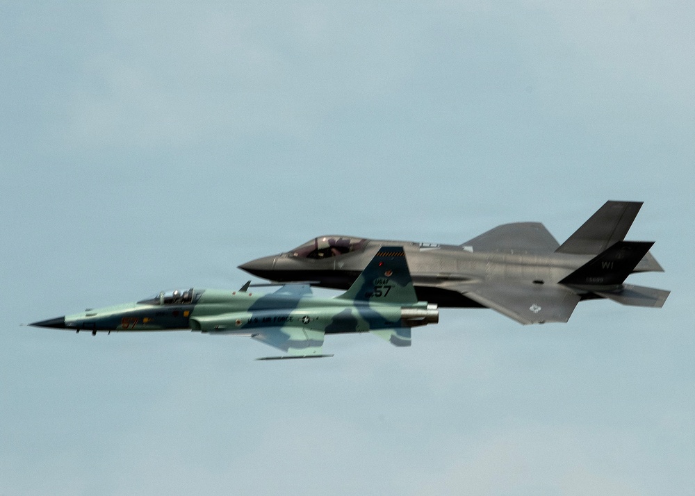 Two U.S. Air Force jet aircraft are flying in close formation against an overcast sky. The foreground aircraft is painted in a green camouflage scheme consistent with a Northrop F-5 aggressor aircraft, while the background aircraft is a modern gray F-35 Lightning II stealth fighter. This appears to be a heritage or demonstration flight pairing an older aggressor-role aircraft with a contemporary fifth-generation fighter.