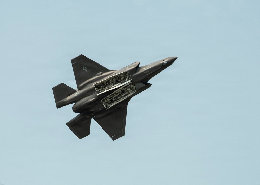 A dark gray stealth fighter jet is photographed in flight against a clear light blue sky, viewed from below at an angle. The aircraft's internal weapons bay doors are open, revealing the internal bay structure and mounted ordnance or pylons. The image captures the aircraft in a banking maneuver, providing a clear view of its underside.