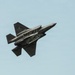 A dark gray stealth fighter jet is photographed in flight against a clear light blue sky, viewed from below at an angle. The aircraft's internal weapons bay doors are open, revealing the internal bay structure and mounted ordnance or pylons. The image captures the aircraft in a banking maneuver, providing a clear view of its underside.