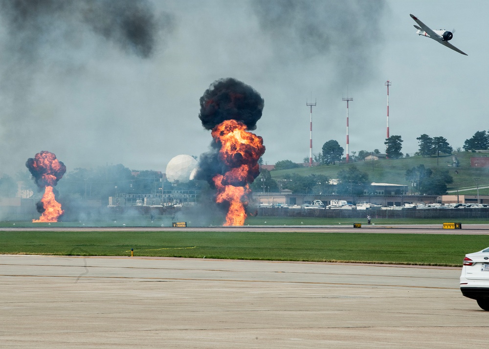 An airshow demonstration at a military or civilian airfield featuring pyrotechnic ground explosions simulating a bombing run. A vintage propeller-driven warbird aircraft is visible in the upper right, flying away from the explosion sequence. The controlled detonations produce large fireballs and black smoke columns along the runway area.