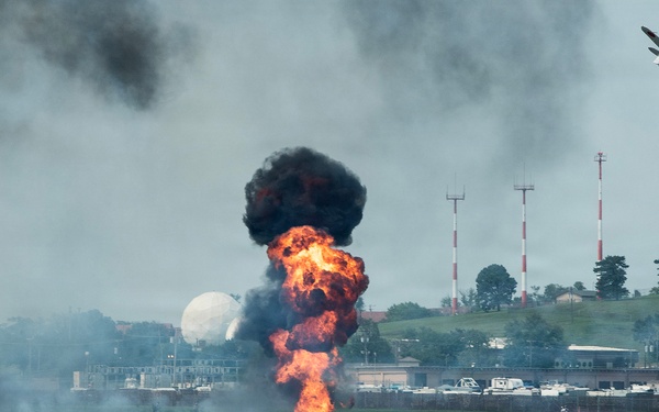An airshow demonstration at a military or civilian airfield featuring pyrotechnic ground explosions simulating a bombing run. A vintage propeller-driven warbird aircraft is visible in the upper right, flying away from the explosion sequence. The controlled detonations produce large fireballs and black smoke columns along the runway area.
