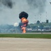 An airshow demonstration at a military or civilian airfield featuring pyrotechnic ground explosions simulating a bombing run. A vintage propeller-driven warbird aircraft is visible in the upper right, flying away from the explosion sequence. The controlled detonations produce large fireballs and black smoke columns along the runway area.