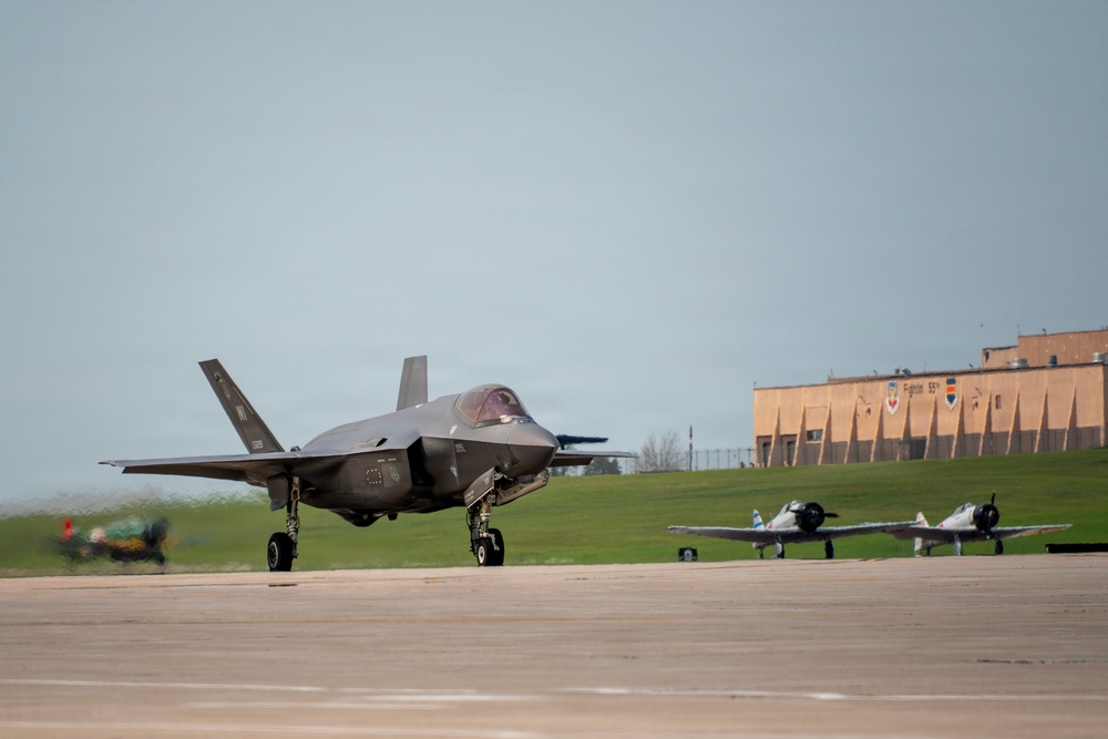 A modern F-35 Lightning II stealth fighter jet is taxiing on a military airbase runway. In the background, two vintage World War II-era propeller aircraft are parked on the tarmac, creating a striking contrast between generations of military aviation. A military hangar building with insignia is visible on a hill in the background.
