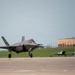 A modern F-35 Lightning II stealth fighter jet is taxiing on a military airbase runway. In the background, two vintage World War II-era propeller aircraft are parked on the tarmac, creating a striking contrast between generations of military aviation. A military hangar building with insignia is visible on a hill in the background.
