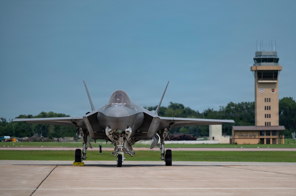 A fifth-generation stealth fighter jet, identifiable as an F-35 Lightning II, is taxiing on a flight line apron with landing gear deployed and gear bay doors open. A military air traffic control tower is visible in the background to the right, with a sign partially readable as 'Offutt' suggesting this may be Offutt Air Force Base in Nebraska. The image is taken from a low, front-facing angle providing a clear view of the aircraft's nose, cockpit, and undercarriage.