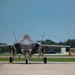 A fifth-generation stealth fighter jet, identifiable as an F-35 Lightning II, is taxiing on a flight line apron with landing gear deployed and gear bay doors open. A military air traffic control tower is visible in the background to the right, with a sign partially readable as 'Offutt' suggesting this may be Offutt Air Force Base in Nebraska. The image is taken from a low, front-facing angle providing a clear view of the aircraft's nose, cockpit, and undercarriage.