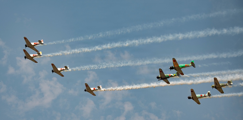 A formation of approximately nine vintage WWII-era Japanese-style aircraft, painted with red rising sun markings, fly in a diagonal echelon formation against a partly cloudy blue sky. Each aircraft trails white smoke, suggesting an airshow or commemorative flight display. One aircraft near the center-right is distinctly painted in green camouflage, differentiating it from the predominantly olive/tan-colored others.