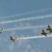 A formation of approximately nine vintage WWII-era Japanese-style aircraft, painted with red rising sun markings, fly in a diagonal echelon formation against a partly cloudy blue sky. Each aircraft trails white smoke, suggesting an airshow or commemorative flight display. One aircraft near the center-right is distinctly painted in green camouflage, differentiating it from the predominantly olive/tan-colored others.