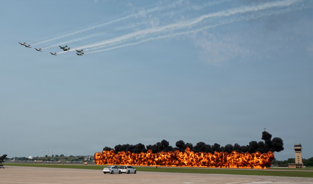 An airshow demonstration featuring a formation of propeller-driven aircraft flying overhead while a large pyrotechnic fire wall burns along the runway below. The controlled explosion produces dramatic orange flames and thick black smoke across a wide stretch of tarmac. A control tower and parked vehicles are visible in the background, confirming this is an active airfield event.