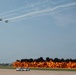 An airshow demonstration featuring a formation of propeller-driven aircraft flying overhead while a large pyrotechnic fire wall burns along the runway below. The controlled explosion produces dramatic orange flames and thick black smoke across a wide stretch of tarmac. A control tower and parked vehicles are visible in the background, confirming this is an active airfield event.