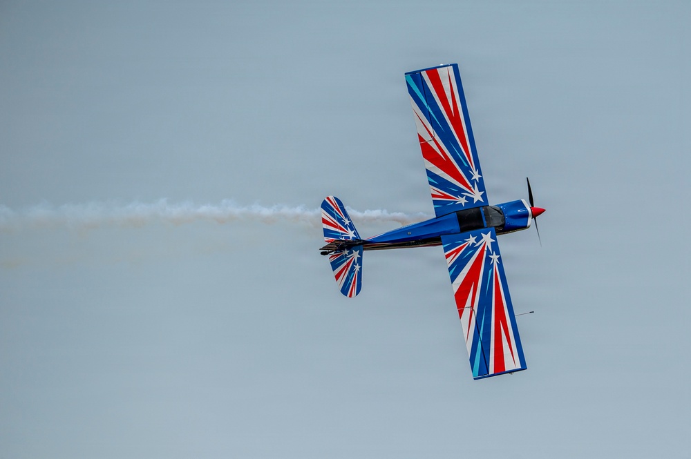 An aerobatic aircraft is captured mid-flight from a below-angle perspective against an overcast sky. The plane features a striking patriotic paint scheme of red, white, and blue with star motifs, and is trailing white smoke, indicating an airshow performance. The aircraft appears to be in a banked or rolling maneuver.