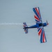 An aerobatic aircraft is captured mid-flight from a below-angle perspective against an overcast sky. The plane features a striking patriotic paint scheme of red, white, and blue with star motifs, and is trailing white smoke, indicating an airshow performance. The aircraft appears to be in a banked or rolling maneuver.