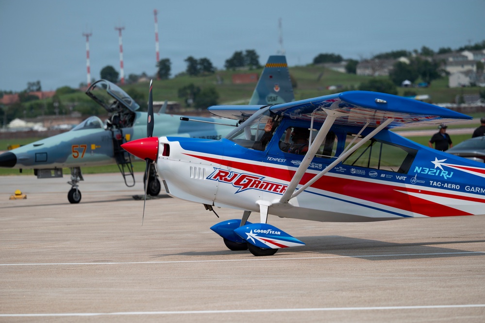 The image shows an airshow tarmac scene featuring a prominently displayed red, white, and blue aerobatic propeller aircraft bearing the name 'RJ Gritter' and registration number N321RJ, sponsored by Goodyear and other companies. Behind it sits a gray military jet fighter marked with the number '57', and further back a USAF aircraft is partially visible. The setting appears to be a military air base or airshow venue with green hills and structures visible in the background.