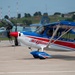 The image shows an airshow tarmac scene featuring a prominently displayed red, white, and blue aerobatic propeller aircraft bearing the name 'RJ Gritter' and registration number N321RJ, sponsored by Goodyear and other companies. Behind it sits a gray military jet fighter marked with the number '57', and further back a USAF aircraft is partially visible. The setting appears to be a military air base or airshow venue with green hills and structures visible in the background.