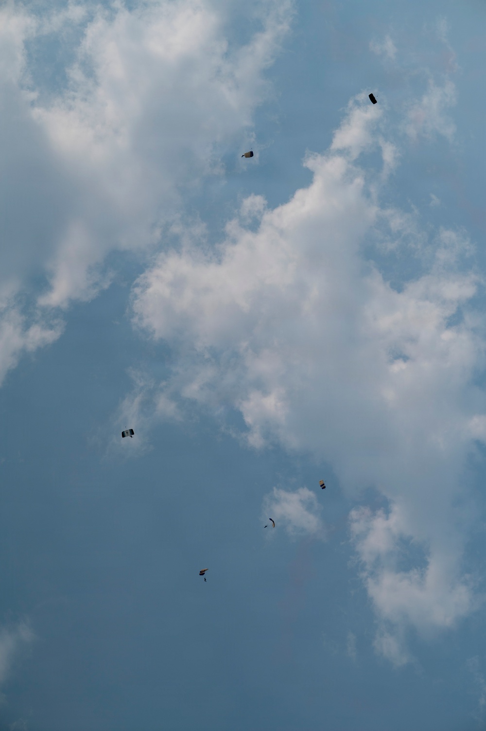 The image shows multiple parachutists descending through a partly cloudy blue sky, photographed from the ground looking upward. At least six parachutes are visible at various altitudes, suggesting a group skydiving or military jump exercise. The subjects are small and distant, indicating they are at significant altitude.
