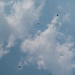 The image shows multiple parachutists descending through a partly cloudy blue sky, photographed from the ground looking upward. At least six parachutes are visible at various altitudes, suggesting a group skydiving or military jump exercise. The subjects are small and distant, indicating they are at significant altitude.