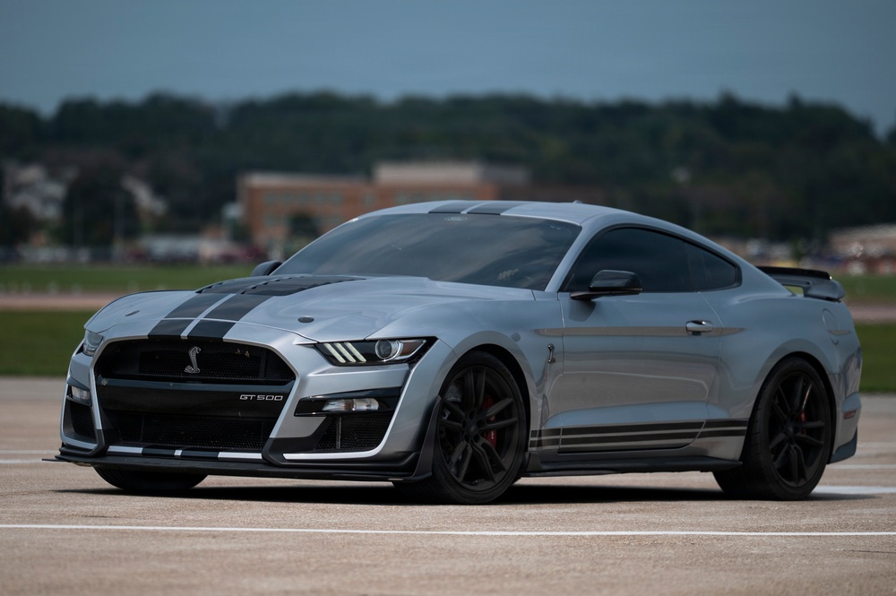 A silver Ford Mustang Shelby GT500 is photographed on what appears to be a tarmac or airfield surface. The car features distinctive dual black racing stripes, a Shelby cobra grille emblem, and red brake calipers visible behind black multi-spoke wheels. The background shows a blurred treeline and buildings under an overcast sky.