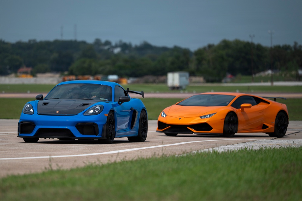 Two high-performance sports cars are positioned side by side on what appears to be a runway or airstrip. On the left is a blue Porsche 718 Cayman GT4 with a carbon fiber hood and rear wing, and on the right is an orange Lamborghini Huracán with a rear spoiler. The setting suggests a track day, drag racing event, or automotive meetup.
