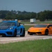 Two high-performance sports cars are positioned side by side on what appears to be a runway or airstrip. On the left is a blue Porsche 718 Cayman GT4 with a carbon fiber hood and rear wing, and on the right is an orange Lamborghini Huracán with a rear spoiler. The setting suggests a track day, drag racing event, or automotive meetup.