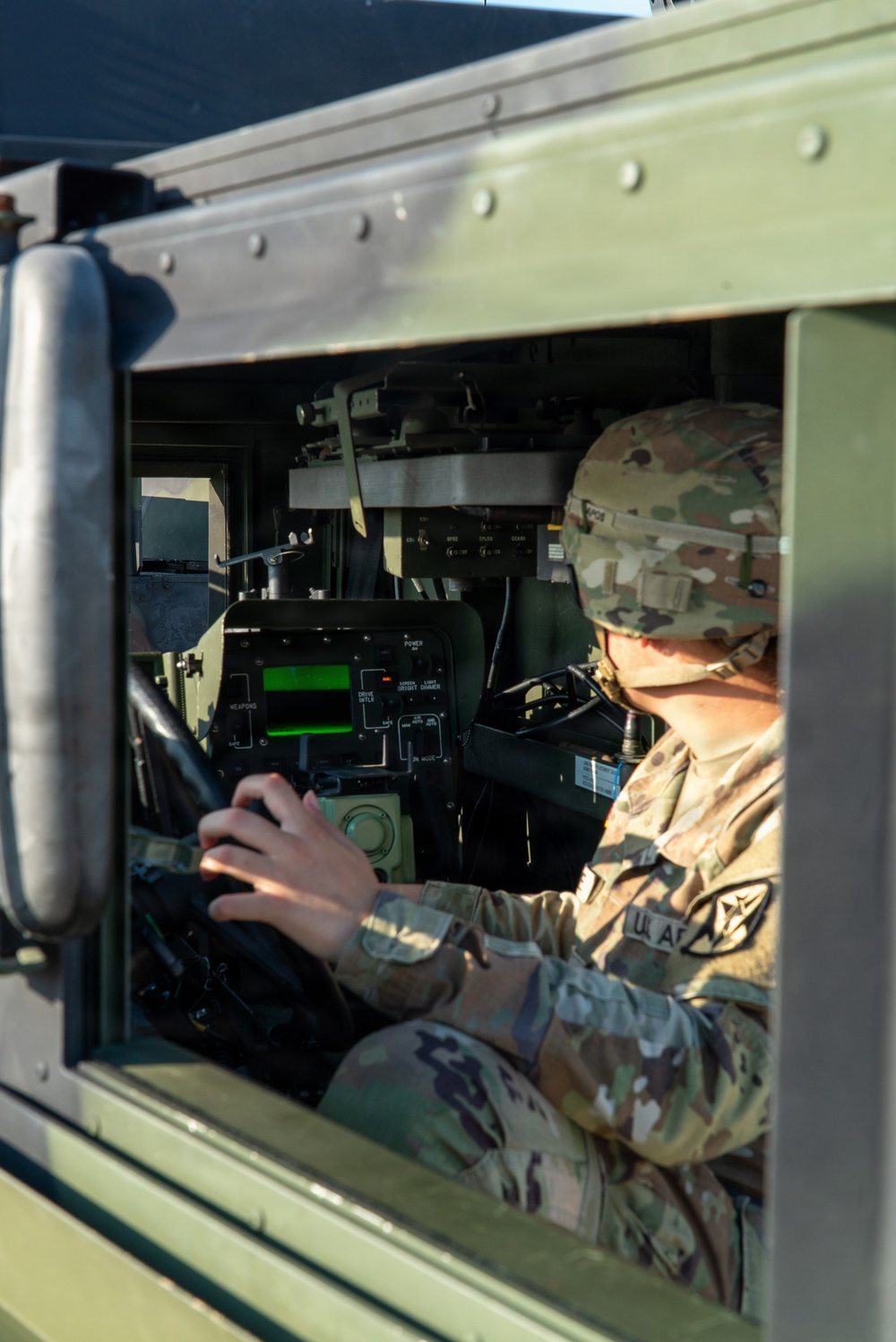 A U.S. Army soldier in Operational Camouflage Pattern (OCP) uniform is seated inside a military vehicle, likely a HMMWV (Humvee), operating interior controls and equipment. The soldier is viewed through the open window frame of the olive-drab colored vehicle. Various military electronic systems and a green-lit display panel are visible on the dashboard.
