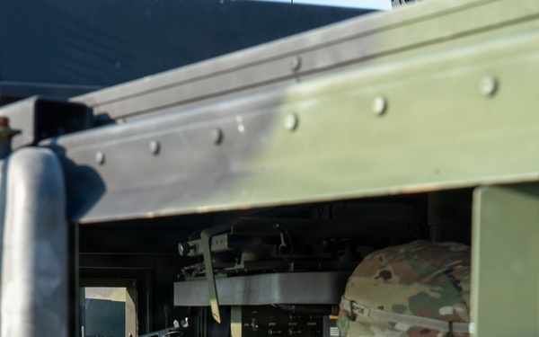 A U.S. Army soldier in Operational Camouflage Pattern (OCP) uniform is seated inside a military vehicle, likely a HMMWV (Humvee), operating interior controls and equipment. The soldier is viewed through the open window frame of the olive-drab colored vehicle. Various military electronic systems and a green-lit display panel are visible on the dashboard.