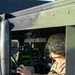 A U.S. Army soldier in Operational Camouflage Pattern (OCP) uniform is seated inside a military vehicle, likely a HMMWV (Humvee), operating interior controls and equipment. The soldier is viewed through the open window frame of the olive-drab colored vehicle. Various military electronic systems and a green-lit display panel are visible on the dashboard.