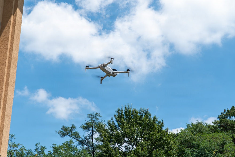 A small consumer quadcopter drone is captured mid-flight against a partly cloudy blue sky. The drone appears to be a light gray/white model with four extended arms and visible rotors in motion. Green trees and the edge of a building are visible in the lower portion of the frame.