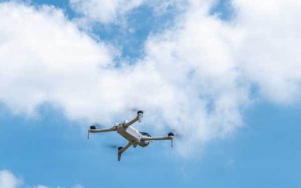A small consumer quadcopter drone is captured mid-flight against a partly cloudy blue sky. The drone appears to be a light gray/white model with four extended arms and visible rotors in motion. Green trees and the edge of a building are visible in the lower portion of the frame.