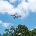 A small consumer quadcopter drone is captured mid-flight against a partly cloudy blue sky. The drone appears to be a light gray/white model with four extended arms and visible rotors in motion. Green trees and the edge of a building are visible in the lower portion of the frame.