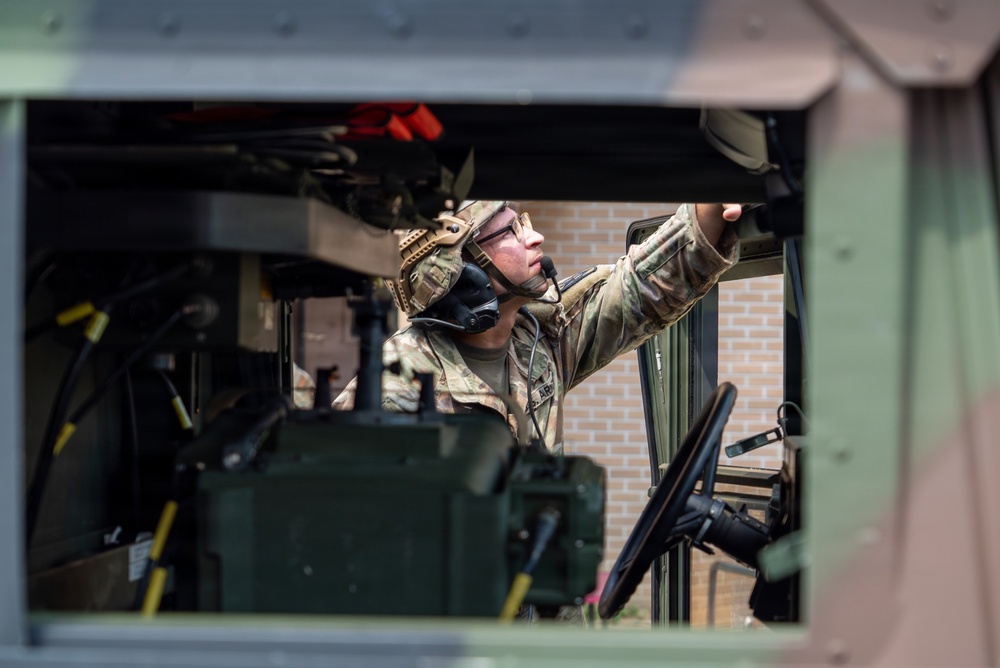 A military service member in full combat gear is performing maintenance or inspection inside a military tactical vehicle, viewed through an open window or hatch. The soldier is wearing multicam camouflage uniform, a tactical helmet, communications headset, and protective eyewear. The image is captured from outside the vehicle looking inward, with a brick building visible in the background.