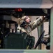 A military service member in full combat gear is performing maintenance or inspection inside a military tactical vehicle, viewed through an open window or hatch. The soldier is wearing multicam camouflage uniform, a tactical helmet, communications headset, and protective eyewear. The image is captured from outside the vehicle looking inward, with a brick building visible in the background.