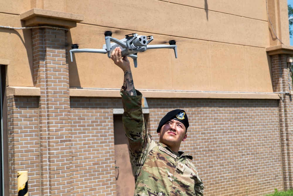A U.S. Air Force service member in OCP (Operational Camouflage Pattern) uniform and black beret is holding a small quadcopter drone aloft with one hand while looking up at it. The name tape on the uniform reads 'COVARRUBIAS' and a unit patch is visible on the left shoulder. The photo is taken outdoors in front of a tan and brick building in what appears to be a military installation.