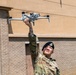 A U.S. Air Force service member in OCP (Operational Camouflage Pattern) uniform and black beret is holding a small quadcopter drone aloft with one hand while looking up at it. The name tape on the uniform reads 'COVARRUBIAS' and a unit patch is visible on the left shoulder. The photo is taken outdoors in front of a tan and brick building in what appears to be a military installation.