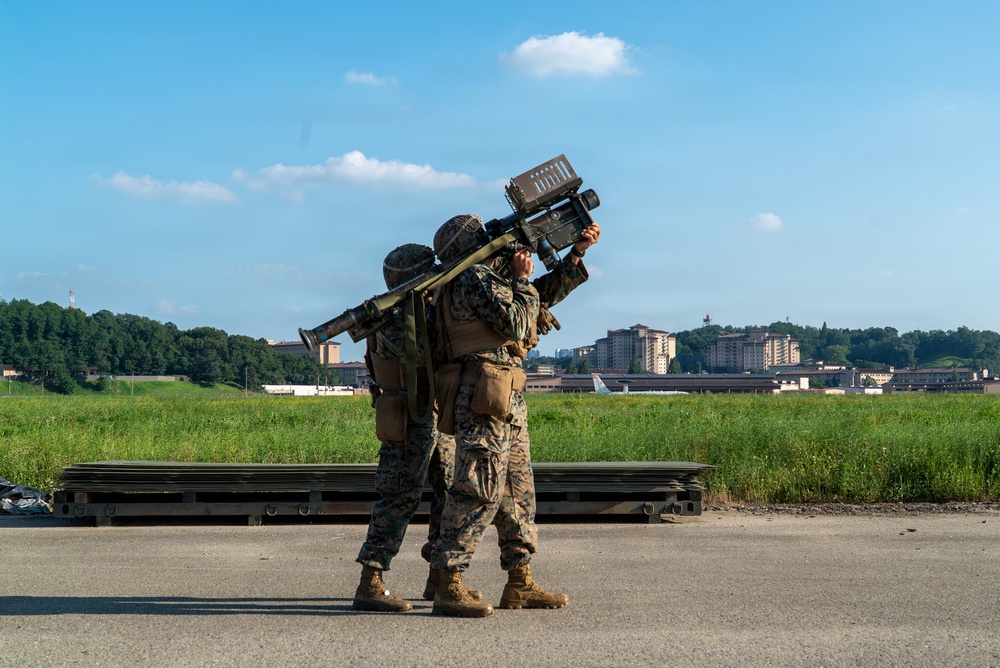 Two military personnel in camouflage uniforms are operating what appears to be a man-portable air-defense system (MANPADS), likely a FIM-92 Stinger or similar shoulder-fired missile system, aimed skyward. They are positioned on a paved surface near what appears to be an airfield or military installation. The background shows green fields, trees, and multi-story buildings consistent with a military base environment.