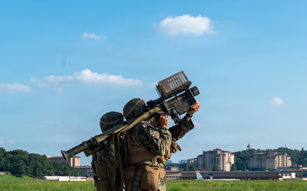Two military personnel in camouflage uniforms are operating what appears to be a man-portable air-defense system (MANPADS), likely a FIM-92 Stinger or similar shoulder-fired missile system, aimed skyward. They are positioned on a paved surface near what appears to be an airfield or military installation. The background shows green fields, trees, and multi-story buildings consistent with a military base environment.