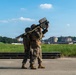 Two military personnel in camouflage uniforms are operating what appears to be a man-portable air-defense system (MANPADS), likely a FIM-92 Stinger or similar shoulder-fired missile system, aimed skyward. They are positioned on a paved surface near what appears to be an airfield or military installation. The background shows green fields, trees, and multi-story buildings consistent with a military base environment.