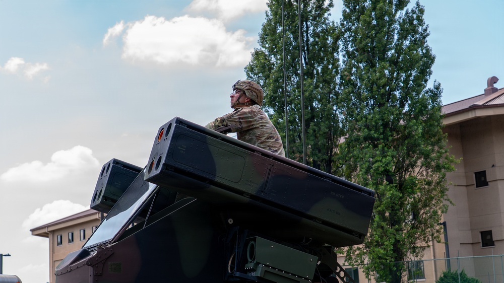 A military service member in camouflage uniform and helmet is positioned atop what appears to be a Avenger air defense system or similar military vehicle-mounted missile launcher. The soldier is looking outward in a watchful posture, suggesting a security or operational role. The setting appears to be a military installation or base, with buildings and trees visible in the background.