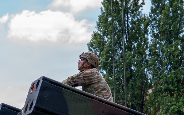 A military service member in camouflage uniform and helmet is positioned atop what appears to be a Avenger air defense system or similar military vehicle-mounted missile launcher. The soldier is looking outward in a watchful posture, suggesting a security or operational role. The setting appears to be a military installation or base, with buildings and trees visible in the background.