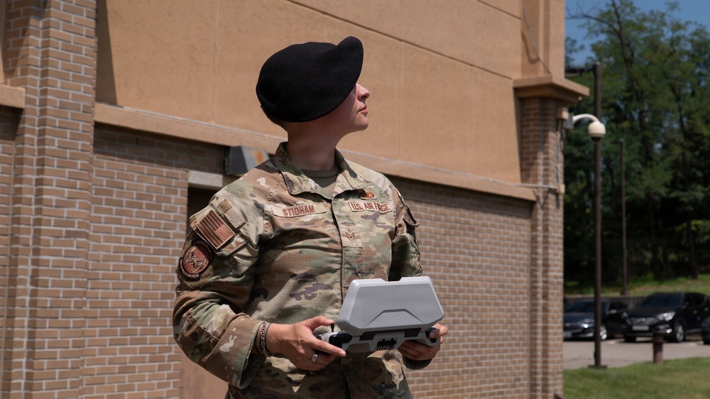 A U.S. Air Force service member wearing OCP (Operational Camouflage Pattern) uniform and black beret is operating what appears to be a drone remote controller outdoors. The name tape reads 'STIDHAM' and the right chest tape reads 'U.S. AIR FORCE.' The individual is looking upward, likely monitoring an airborne drone, while standing near a brick building on what appears to be a military installation.