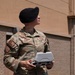 A U.S. Air Force service member wearing OCP (Operational Camouflage Pattern) uniform and black beret is operating what appears to be a drone remote controller outdoors. The name tape reads 'STIDHAM' and the right chest tape reads 'U.S. AIR FORCE.' The individual is looking upward, likely monitoring an airborne drone, while standing near a brick building on what appears to be a military installation.