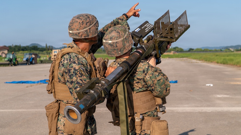 Two military personnel in MARPAT (Marine pattern) camouflage uniforms are operating what appears to be a man-portable air-defense system (MANPADS), likely a FIM-92 Stinger missile system, on a paved surface such as an airfield or tarmac. One soldier is pointing upward, possibly tracking or designating an aerial target, while the other shoulders the launcher. The setting appears to be a training exercise based on the controlled environment and visible background infrastructure.