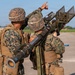 Two military personnel in MARPAT (Marine pattern) camouflage uniforms are operating what appears to be a man-portable air-defense system (MANPADS), likely a FIM-92 Stinger missile system, on a paved surface such as an airfield or tarmac. One soldier is pointing upward, possibly tracking or designating an aerial target, while the other shoulders the launcher. The setting appears to be a training exercise based on the controlled environment and visible background infrastructure.