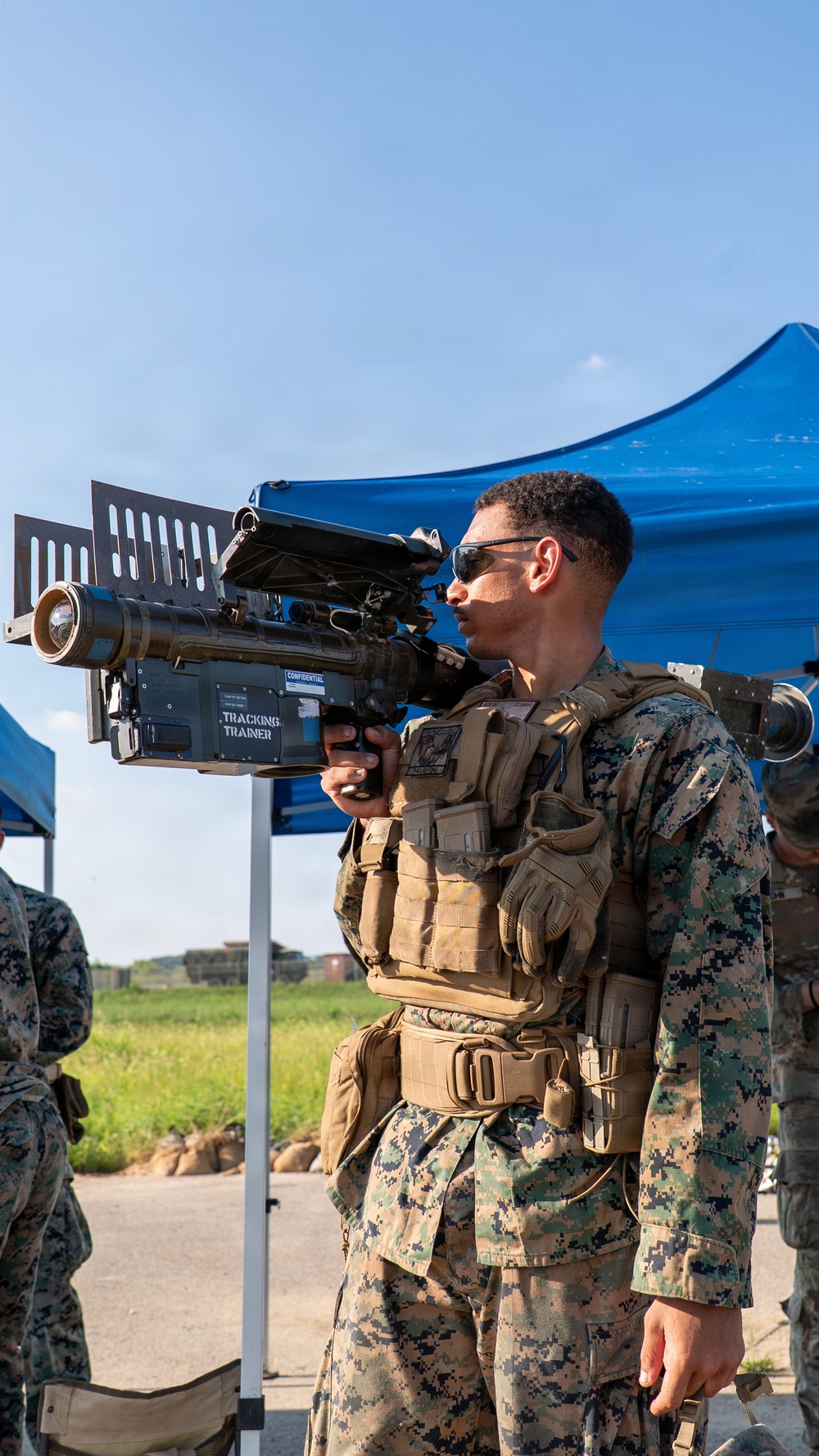 A U.S. Marine in MARPAT digital camouflage uniform and full tactical gear is holding and looking through a shoulder-fired missile tracking trainer device labeled 'TRACKING TRAINER' and marked 'CONFIDENTIAL'. The setting appears to be an outdoor training exercise or demonstration area, with a blue canopy tent visible in the background. Additional Marines in similar uniforms are partially visible in the background.
