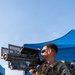 A U.S. Marine in MARPAT digital camouflage uniform and full tactical gear is holding and looking through a shoulder-fired missile tracking trainer device labeled 'TRACKING TRAINER' and marked 'CONFIDENTIAL'. The setting appears to be an outdoor training exercise or demonstration area, with a blue canopy tent visible in the background. Additional Marines in similar uniforms are partially visible in the background.